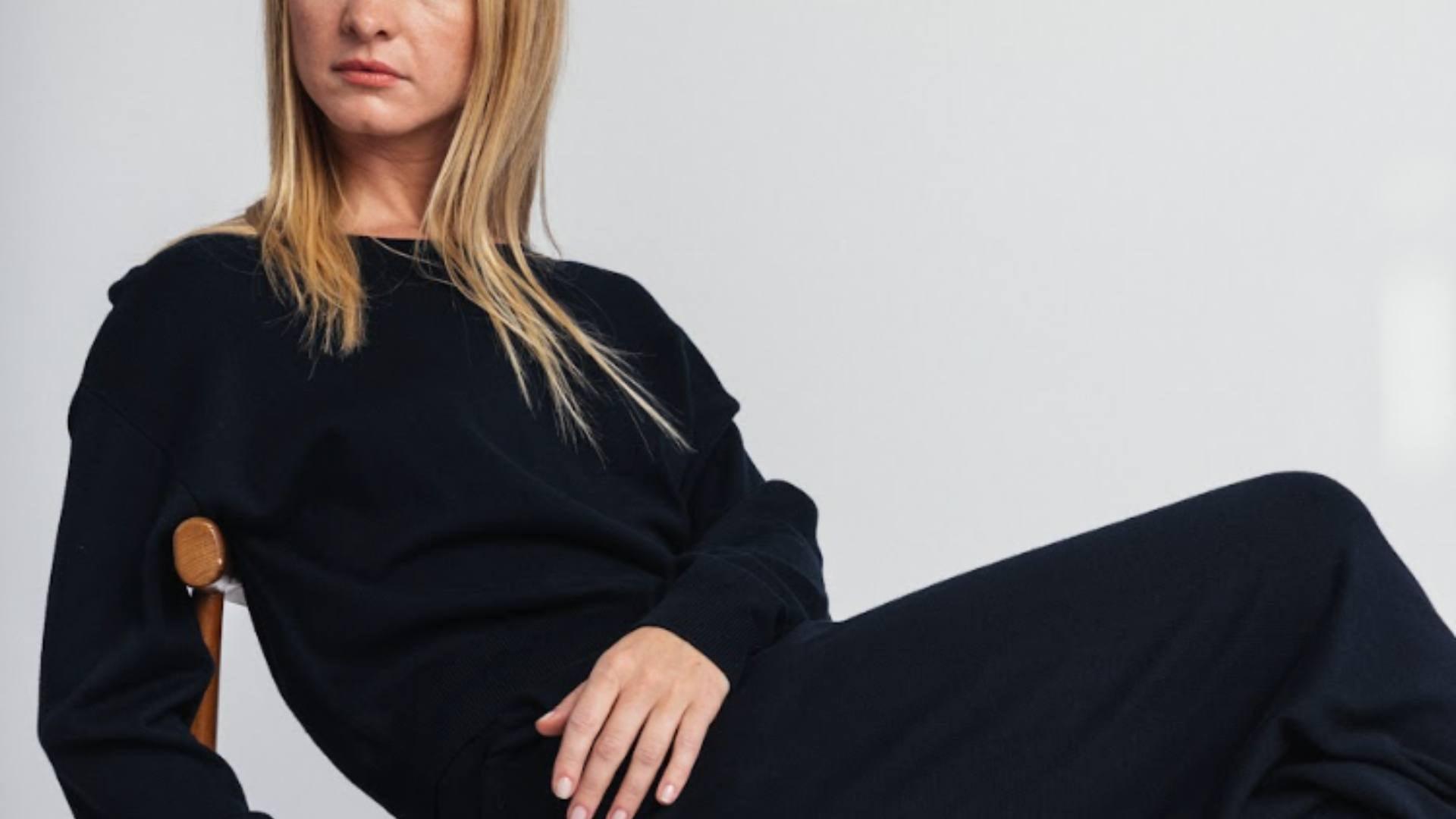 A blonde woman sits on a wooden chair and poses in a black, sustainable, conscious outfit.