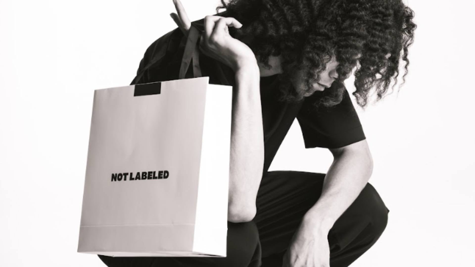A black and white photo shows a man kneeling while looking off to the side while holding a white Not Labeled shopping bag, demonstrating ethical fashion options.