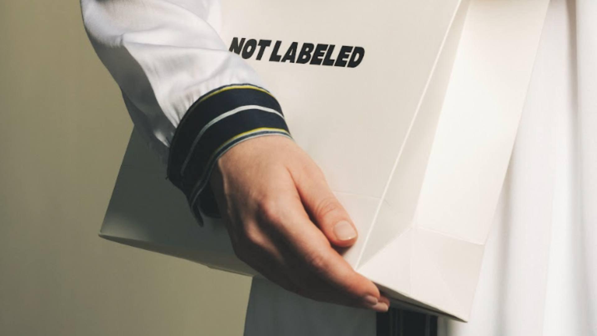 A person wears a white jacket with black and white striped cuffs while holding a Not Labeled gift bag.
