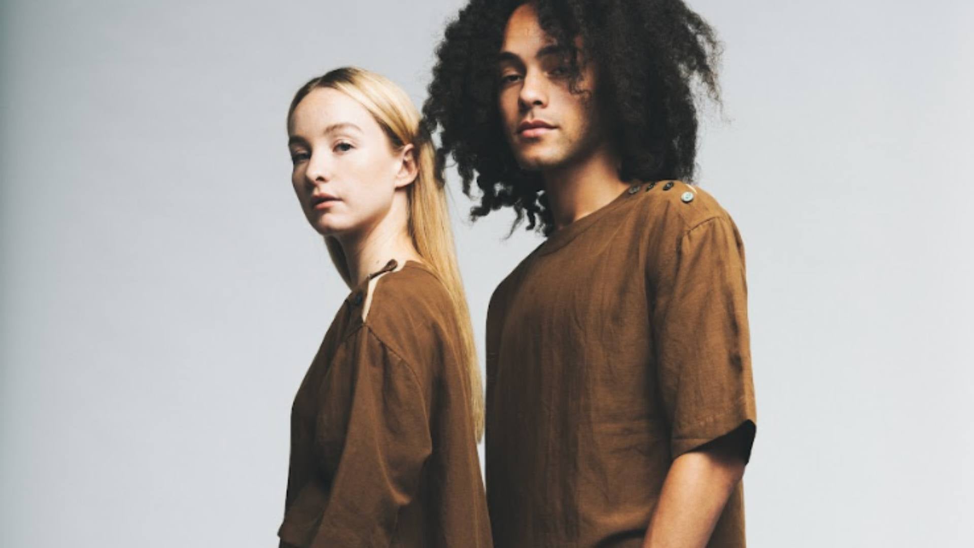 One blonde woman and one brunette man stand together and look at the camera while wearing matching brown shirts, demonstrating sustainable Valentine's Day options.
